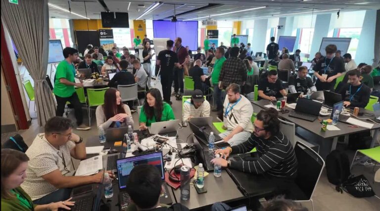 Large group of people working collaboratively on laptops at tables in a brightly lit room, likely participating in a hackathon or workshop event.
